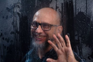 Islak camın arkasında gözlüklü sakallı kel bir adamın komik hareketler yaptığı yakın plan portresi. Siyah arkaplanda izole.