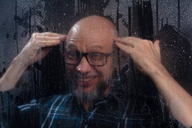 Islak camın arkasında gözlüklü sakallı kel bir adamın komik hareketler yaptığı yakın plan portresi. Siyah arkaplanda izole.