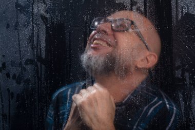 Islak camın arkasında gözlüklü sakallı kel bir adamın komik hareketler yaptığı yakın plan portresi. Siyah arkaplanda izole.