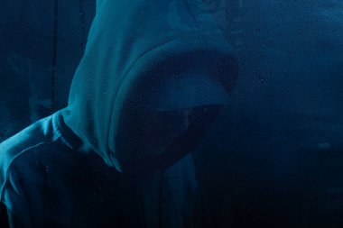 Islak camın arkasında sweatshirt giyen gizemli bir adamın portresi. Yüzü gizli. Stüdyo portresi. Mavi arkaplanda izole.