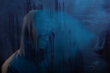 Islak camın arkasında sweatshirt giyen gizemli bir adamın portresi. Yüzü gizli. Stüdyo portresi. Mavi arkaplanda izole.