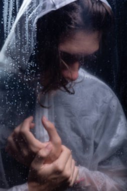 Islak bardağın arkasında elini tutan yağmurluk giyen genç bir adam. Stüdyo portresi. Koyu arkaplanda izole.