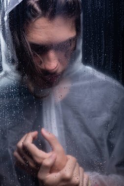 Islak bardağın arkasında elini tutan yağmurluk giyen genç bir adam. Stüdyo portresi. Koyu arkaplanda izole.