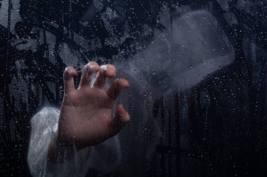 Islak camın arkasında yağmurluk giyen insan portresi. Elinle cama dokunuyorsun. Stüdyo portresi. Koyu arkaplanda izole.