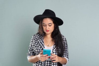 Stüdyoda cep telefonuna bakan genç ve güzel bir kadın. Açık yeşil arka planda siyah şapka takıyor..