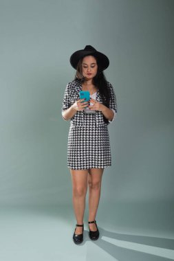 Stüdyoda cep telefonuna bakan genç ve güzel bir kadın. Açık yeşil arka planda siyah şapka takıyor..