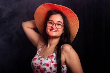 Plaj şapkası takan ve fotoğraf stüdyosunda poz veren siyah saçlı güzel bir genç kadın portresi. Koyu dumanlı arkaplanda izole.