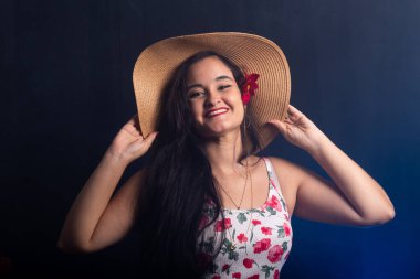 Siyah saçlı, güzel, gülümseyen bir genç kadın kahverengi bir plaj şapkası takıyor ve sol kulağında kırmızı bir çiçek var. Renkli giysiler içinde kameraya bakıyor. Koyu dumanlı arkaplanda izole.