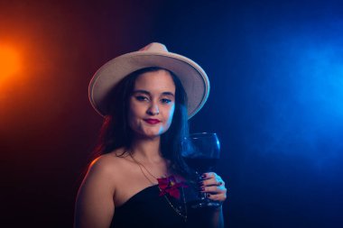 Kahverengi şapkalı güzel bir kadın elinde bir bardak şarapla kameraya bakıyor. Stüdyo fotoğrafı dumanlı renkli arkaplan.