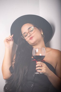 Siyah kıyafetli güzel bir kadın portresi ve duvara yaslanmış bir bardak şarap taşıyan şapkalı fotoğraf için poz veren. Beyaz stüdyo duvarında izole edilmiş..