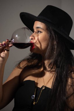 Siyah elbiseli, şapkalı, cam bardaktan şarap içen, duvarda gölgeler bırakan güzel bir kadın. Stüdyo fotoğrafı
