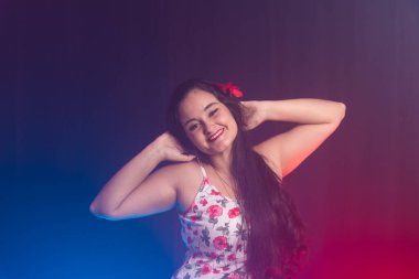 Renkli elbiseler giyen, sol kulağında kırmızı bir çiçek olan güzel siyah saçlı kadın fotoğraf için poz veriyor. Renkli dumanlı arkaplanda izole.