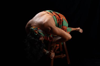 Tahta taburede oturan, renkli kıyafetler giyen, vücut akrobasisi yapan güzel olgun bir kadının portresi. Siyah arkaplanda izole.