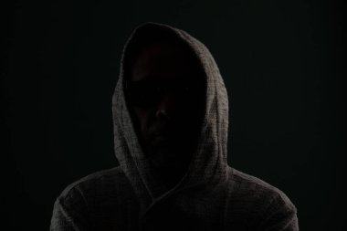 Güneş gözlüğü takan ve başını örten kapüşonlu kazak giyen gizemli adamın silueti. Koyu arkaplanda izole.