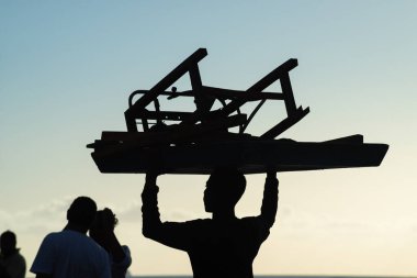 Salvador, Bahia, Brezilya - 21 Aralık 2019: Bir plaj işçisi gün batımında siluette görüldü, kafasında Salvador, Bahia 'da tahta sandalyeler vardı..