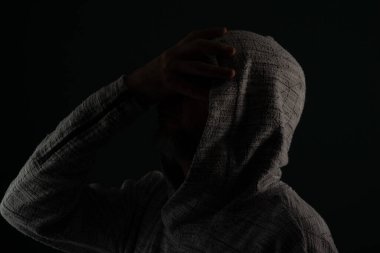 Güneş gözlüğü takan ve kafasında elleri olan kapüşonlu kazak giyen gizemli bir adamın portresini yakından çek. Koyu arkaplanda izole.