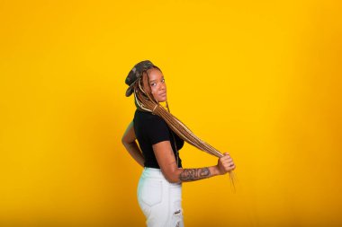 Saçında örgüleri olan, üzerinde pantolon ve bluzla kameraya poz veren güzel bir genç kadının stüdyo portresi. Sarı arkaplanda izole edilmiş.