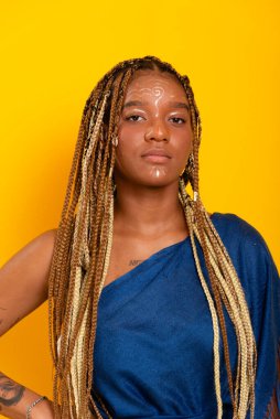 Saçları örgülü, mavi elbiseli ve kameraya bakan güzel bir genç kadın. Sarı arkaplanda izole edilmiş.