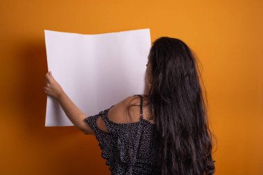 Güzel siyah saçlı Latin Amerikalı kadın kameraya arkası dönük, ayakta duran boş bir pankart tutuyor. Hardal renginde arkaplanda izole.