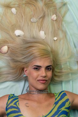 Güzel sarışın bir kadın açık yeşil bir kanepede uzanmış, saçlarında deniz kabukları, kameraya bakıyor. Salvador, Bahia.