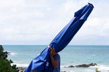 Salvador, Bahia, Brezilya - 11 Ekim 2019: Bahia 'nın Salvador kentindeki Barra plajında güçlü güneş ışığı altında bir plaj işçisi mavi bir şemsiye taşırken görüldü..