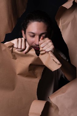 A man peering through a hole in a sheet of brown paper. Tearing the paper with his hands. Studio portrait. clipart