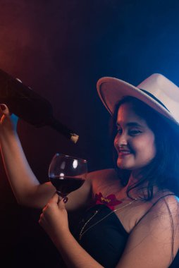 Kahverengi şapkalı ve siyah elbiseli güzel bir kadın bir şişe ve cam kadehle şarap doldurma simülasyonu yapıyor. Stüdyo fotoğrafı dumanlı renkli arkaplan.