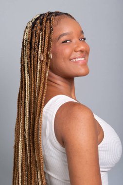 Saçları örgülü, kot pantolon ve beyaz gömlek giyen, kameraya bakıp gülümseyen güzel esmer kadın. Gri stüdyo arka planında izole edilmiş..