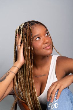 Saçında örgüleri olan güzel esmer bir kadın stüdyo zemininde oturuyor, tahta bir tabureye yaslanıyor, saçını tarıyor. Sakin ve kendinden emin biri. Gri arkaplanda izole.