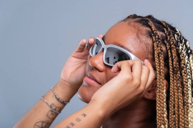 Saçında örgüleri olan güzel bir esmer, yüzünde güneş gözlüğü olan, odaklanmış, kot pantolon ve beyaz bir bluz giyen. Neşeli ve kendine güvenen biri. Gri arkaplanda izole.