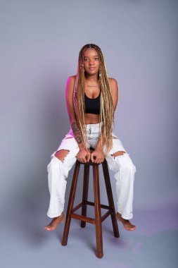 Saçında örgüler, beyaz kot pantolon ve siyah bluz giyen esmer bir manken, tahta bir taburede kameraya poz veriyor. Gri bir arkaplanda izole edilmiş.