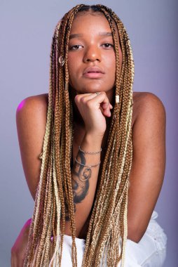 Saçında örgüler, beyaz kot pantolon ve siyah bluz giyen esmer bir manken, tahta bir taburede kameraya poz veriyor. Gri bir arkaplanda izole edilmiş.