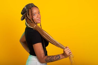 Saçında örgüleri olan, üzerinde pantolon ve bluzla kameraya poz veren güzel bir genç kadının stüdyo portresi. Sarı arkaplanda izole edilmiş.