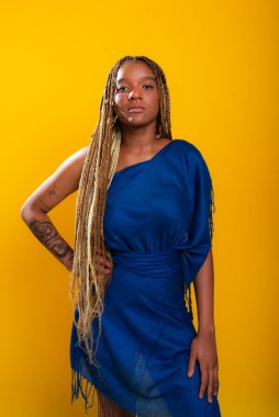 Saçları örgülü, mavi elbiseli ve kameraya bakan güzel bir genç kadın. Sarı arkaplanda izole edilmiş.