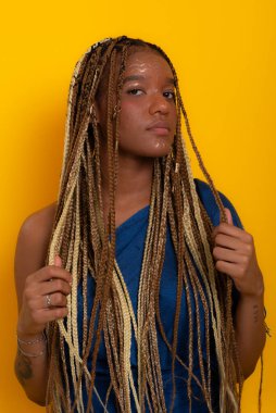 Mavi elbiseli, saçlarını ören ve kameraya poz veren güzel esmer bir kadının fotoğrafı. Sarı arkaplanda izole edilmiş.