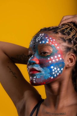 Siyah bluz giyen, saçları örgülü, yüzü renkli boyalı, konsantre bir kadın. Sarı arkaplanda izole edilmiş.