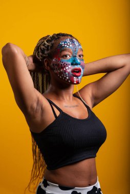 Fotoğraf çekimi sırasında yüzünde renkli boya olan ve örgülerine dokunan genç ve güzel bir kadın. Sarı arkaplanda izole edilmiş.