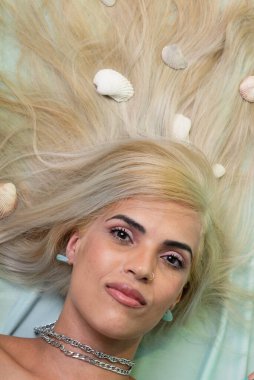 Güzel sarışın bir kadın açık yeşil bir kanepede uzanmış, saçlarında deniz kabukları, kameraya bakıyor. Salvador, Bahia.