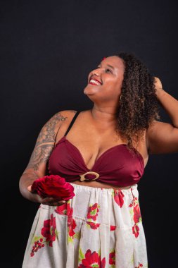 Salvador, Bahia, Brezilya - 21 Ekim 2024: Geleneksel kıyafetler giyen, elinde kırmızı bir gül tutan ve kameraya poz veren güzel siyah kadın. Siyah arkaplanda izole.