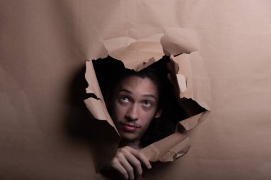 Adam yüzü ve başı dışarı çıkmış ve kahverengi kağıttaki delikten dışarı bakıyor. Eller kağıdı kenara itiyor. Stüdyo resmi.