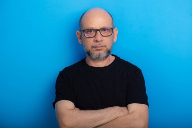Sakallı, kel, reçeteli gözlüklü, kolları çaprazlanmış, mavi arka planda fotoğraf için poz veren bir adam. Stüdyo çekimi.