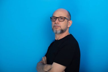 Sakallı, kel, reçeteli gözlüklü, kolları çaprazlanmış, mavi arka planda fotoğraf için poz veren bir adam. Stüdyo çekimi.