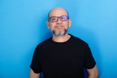 Sakallı, kel, reçeteli gözlüklü ve siyah gömlekli bir adam mavi arka planda fotoğraf için poz veriyor. Stüdyo çekimi.