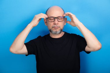 Sakallı, kel, reçeteli gözlük takan ve kolları kameraya dönük siyah gömlekli bir adam. Kendinden emin ve sakin. Stüdyo çekimi.