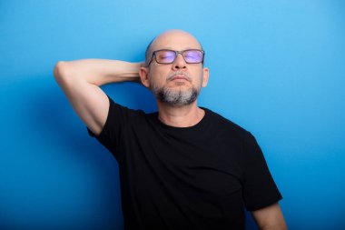 Sakallı, kel, gözlüklü ve tek kollu siyah gömlekli bir adam. Kendinden emin ve odaklanmış. Stüdyo çekimi.