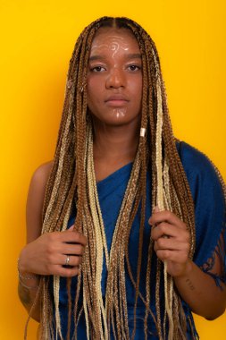 Mavi elbiseli, saçlarını ören ve kameraya poz veren güzel esmer bir kadının fotoğrafı. Sarı arkaplanda izole edilmiş.