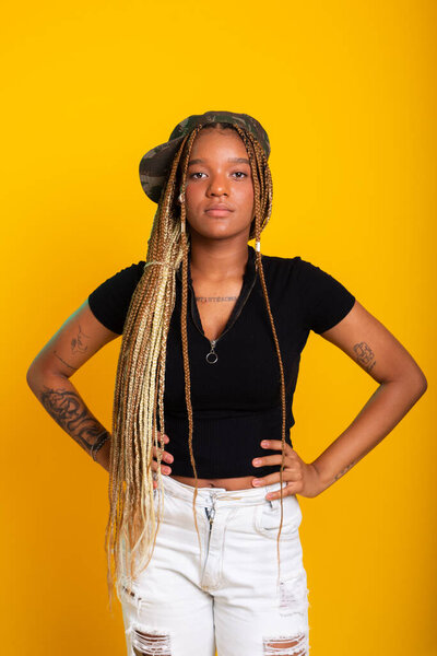 Studio portrait of beautiful young woman with braids in her hair, standing wearing pants and blouse with green hat posing for the camera. Isolated on yellow background.