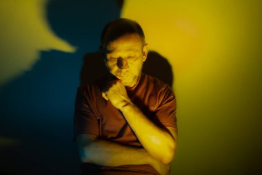 Beyaz adam, üzerinde mavi ve sarı renkli ışıklar olan bir duvara yaslanıyor. Aşağıya bakıyorum. Stüdyo portresi. Flaş sanat.