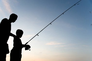 Salvador, Bahia, Brazil - November 16, 2019: Man and child, in Silhouette, are seen fishing during sunset in the city of Salvador, Bahia. clipart