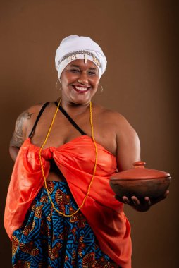 Salvador, Bahia, Brezilya - 21 Ekim 2024: Kahverengi arka planda kilden bir tencere tutan geleneksel Candomble elbiseli güzel bir kadın. Salvador, Bahia.